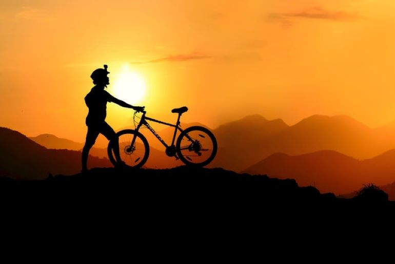 There is a person with a bike at the top of a hill, during the sunset.